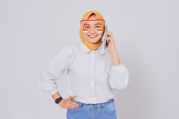 Smiling happy young Asian Muslim woman wearing a hijab talking on mobile phone and looking at camera isolated over white background. Celebrate Indonesian independence day on 17 August