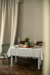 vintage white table with white tablecloth in a room with beige walls and antique armchairs. decorated table with appetizers, bread, vegetables, berries and a vase of daisies.