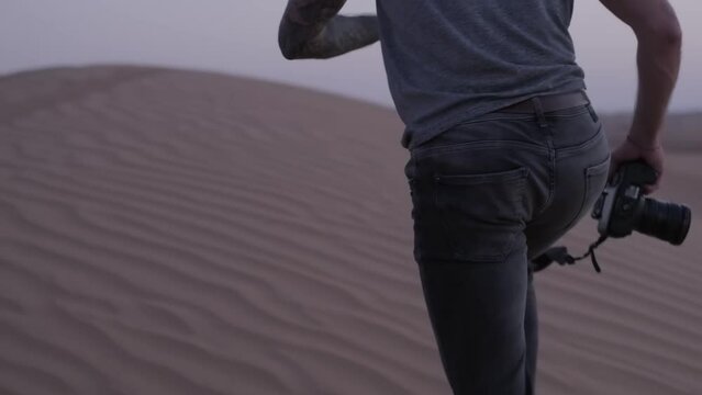 A young photographer with a camera in his hands walks barefoot through the sand dunes of the desert at sunset