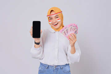 Smiling Happy young Asian Muslim woman holding cash money and showing display mobile phone blank...