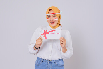 Excited young Asian Muslim woman wearing a hijab holding a gift voucher certificate isolated over...