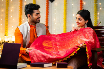 Happy smiling indian wife showing saree design to husband during diwali festival celebration at home to wear - concept of togetherness, excitement and festive preparation