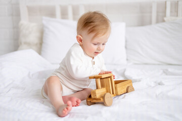 smiling baby 6 months old blond boy is sitting on a large bed in a bright bedroom and playing with a wooden toy car in a cotton bodysuit, the concept of children's goods