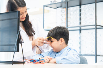 Young optometrist woman or doctor helping Indian cute child boy have fun trying and choosing a new eyeglasses frame at ophthalmology clinic or optics store, smiling. Selective focus on boy