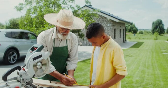 An elderly man in an apron and hat teaches his grandson how to use a miter saw while standing in the backyard of his house