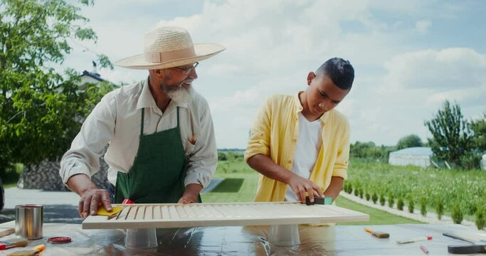 A grandfather in an apron and hat teaches his grandson to sandpaper wood, standing in the backyard on a summer day