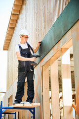 Carpenter constructing wooden framed house. Man worker in glasses cladding facade of house with...