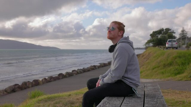 A video of a man wearing son glasses on a bench. The bench is located at a beach in Paraparaumu New Zealand.