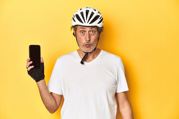 Cyclist man showing phone on yellow backdrop shrugs shoulders and open eyes confused.