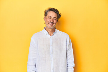 Middle-aged man posing on a yellow backdrop laughs and closes eyes, feels relaxed and happy.