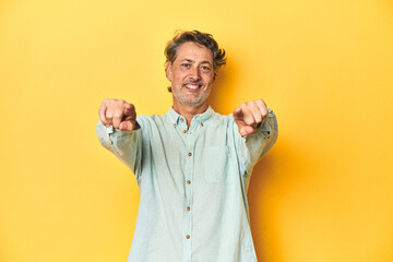 Middle-aged man posing on a yellow backdrop cheerful smiles pointing to front.