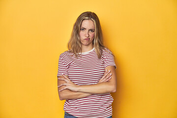 Young blonde Caucasian woman in a red striped t-shirt on a yellow background, frowning face in displeasure, keeps arms folded.