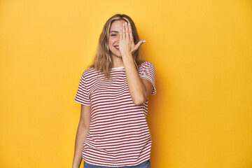 Young blonde Caucasian woman in a red striped t-shirt on a yellow background, having fun covering half of face with palm.