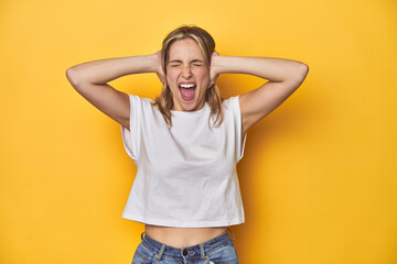 Young blonde Caucasian woman in a white t-shirt on a yellow studio background, covering ears with...