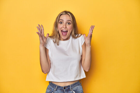 Young blonde Caucasian woman in a white t-shirt on a yellow studio background, celebrating a victory or success, he is surprised and shocked.