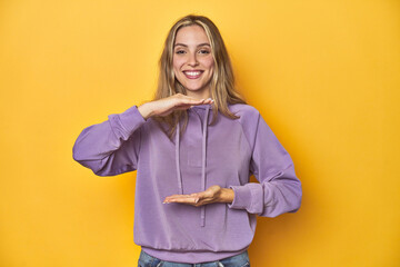 Young blonde Caucasian woman in a violet sweatshirt on a yellow background, holding something with...