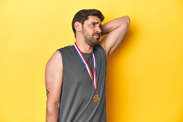 Athletic man with gold medal, yellow studio backdrop touching back of head, thinking and making a...