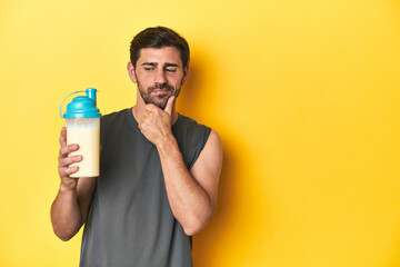 Fit man with protein shake, yellow studio background looking sideways with doubtful and skeptical...