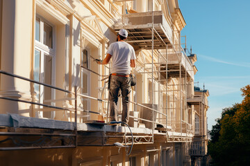 Renovation, restoration, refurbishment. Unrecognizable worker renovating wall of classical style building, standing on scaffolding. Construction worker prepares house facade wall for painting outdoors - 624297843