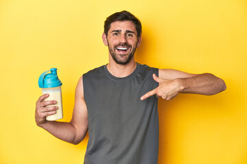 Fit man with protein shake, yellow studio background person pointing by hand to a shirt copy space,...