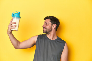 Fit man with protein shake, yellow studio background touching back of head, thinking and making a...