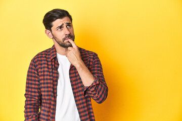 Caucasian man in red checkered shirt, yellow backdrop looking sideways with doubtful and skeptical...