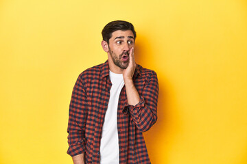 Caucasian man in red checkered shirt, yellow backdrop is saying a secret hot braking news and looking aside