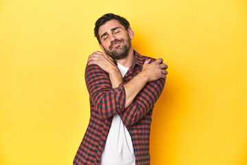 Caucasian man in red checkered shirt, yellow backdrop hugs, smiling carefree and happy.