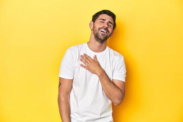 Caucasian man in white t-shirt on yellow studio background laughs out loudly keeping hand on chest.