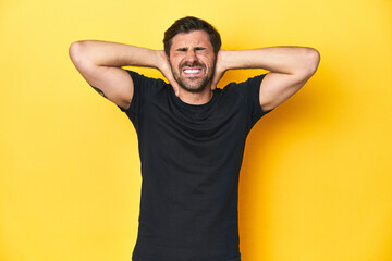 Caucasian man in black t-shirt, yellow studio backdrop covering ears with hands trying not to hear too loud sound.