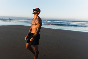 Chico joven tatuado y musculoso sonriendo y posando en playa paradisiaca de andalucia al amanecer