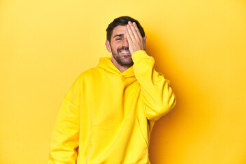 Man in yellow hoodie, monochrome studio backdrop having fun covering half of face with palm.
