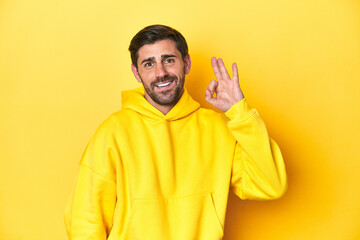Man in yellow hoodie, monochrome studio backdrop cheerful and confident showing ok gesture.