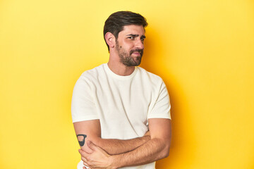 Caucasian man in white t-shirt on yellow studio background suspicious, uncertain, examining you.