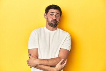 Caucasian man in white t-shirt on yellow studio background blows cheeks, has tired expression. Facial expression concept.