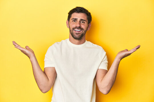 Caucasian man in white t-shirt on yellow studio background makes scale with arms, feels happy and confident.