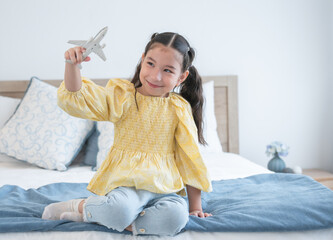 Cute Caucasian little girl with pony tails hair sitting on bed at home and playing with airplane...