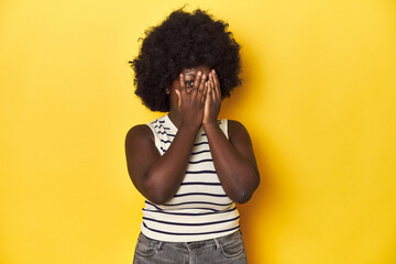 Fototapeta na wymiar African-American woman with afro, studio yellow background blink through fingers frightened and nervous.