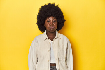 Fototapeta na wymiar African-American woman with afro, studio yellow background blows cheeks, has tired expression. Facial expression concept.