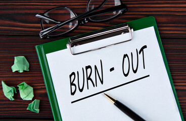BURN-OUT - words on a white sheet on a brown wooden background with a pen and glasses