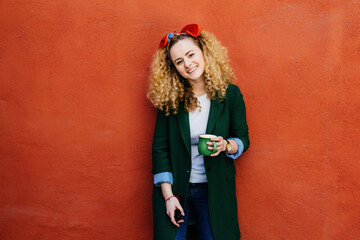 Fashionable Caucasian woman with curly hair, headband, and stylish jacket, holding a green cup of...