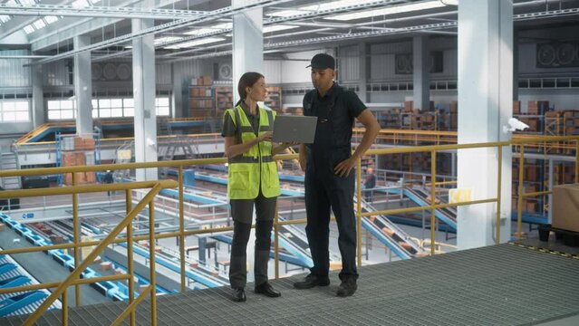 Caucasian Female Manager Talking to Multiethnic Male Distribution Specialist And Using Laptop Computer In Modern Warehouse Facility With Conveyor Belt. Boxes Being Packaged And Delivered To Clients.