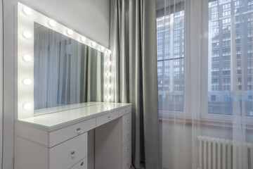 White dressing table in the bedroom interior. The mirror is illuminated. A window with .gray curtains.