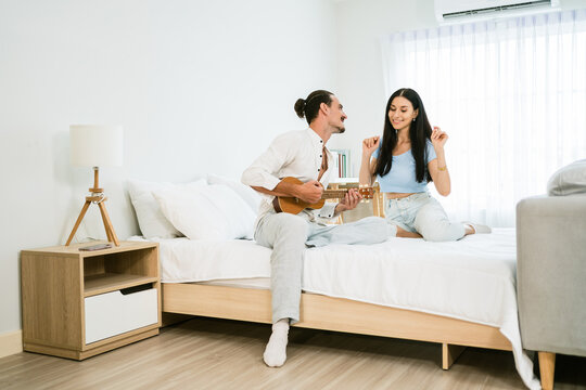 Loving Caucasian couple enjoying the music together. A man is playing the ukulele sitting with a woman on the bed together in a happy mood in the bedroom. Image with copy space.
