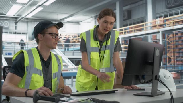 Caucasian Male Stocking Associate Talking to Female Manager And Using Desktop Computer In Warehouse Facility With Automated Conveyor Belt. Man And Woman Working In Sorting Center of Online Marketplace
