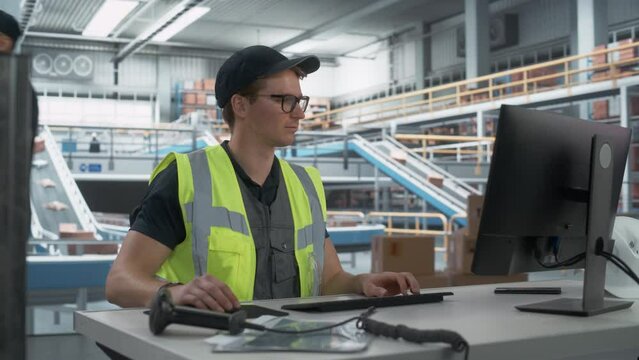 Caucasian Male Stocking Associate Checking Inventory On Desktop Computer In Modern Warehouse Facility With Automated Conveyor Belt. Employees Carrying Boxes to Package Products And Deliver to Clients.