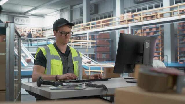 Male Stocking Associate Checking Inventory On Desktop Computer In Warehouse Facility With Automated Conveyor Belt. Sorting Center Employees Carrying Boxes to Package Products And Deliver to Clients.