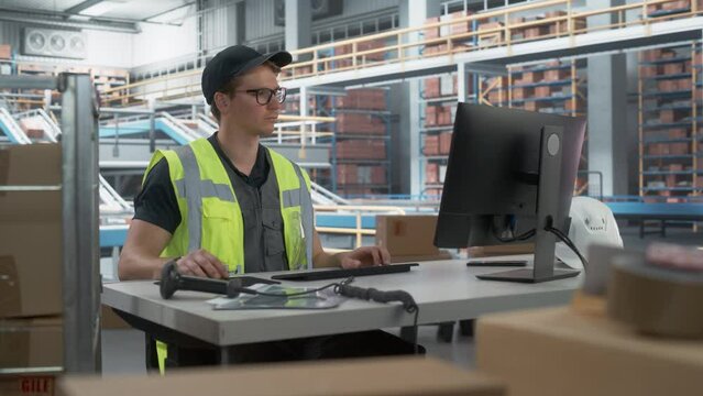 Male Stocking Associate Checking Inventory On Desktop Computer In Warehouse Facility With Automated Conveyor Belt. Logistics Company Employees Carrying Boxes to Package Products And Deliver to Clients