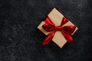 A box wrapped in craft paper and tied with a red ribbon with a bow on a black table, from above....