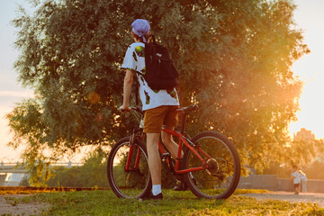 A young man stands with a bicycle on the grass in a city park at sunset.